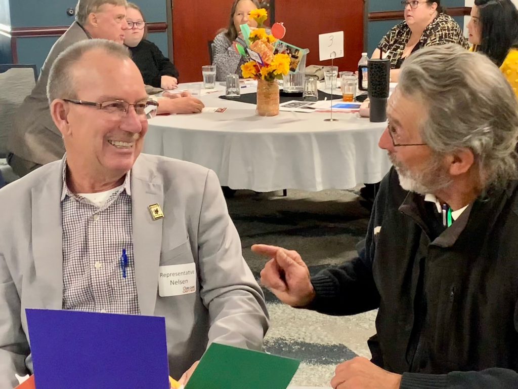 Representative Nelson from Idaho District 26 speaking with provider Bill Benkula from Twin Falls. In the background a group of advocates sitting around table speaking to Idaho Representative Gannon from District 17.